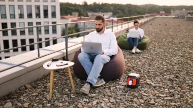 Fasulye koltukta oturan işadamı kabloyu evin çatısındaki taşınabilir elektrik santraline bağlıyor. Arka planda dizüstü bilgisayarda çalışan başka biri.