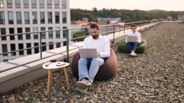 Çatıda dizüstü bilgisayarlar ve şehir ortamında fasulye torbalarıyla çalışan iki kişi. Şehir manzaralı sıradan bir çalışma alanı. Uzaktan çalışma, teknoloji, rahatlama ve üretkenlik kavramı.