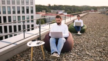 Çatıdaki koltuklarda dizüstü bilgisayarlarla çalışan iki kişi. Arka planda şehir manzarası olan rahat bir çalışma ortamı. Ön plandaki adam düşünceli görünüyor..