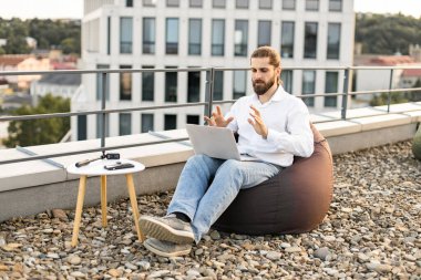 Gündelik kıyafetli bir adam dışarıda dizüstü bilgisayarla çalışıyor. Kişi çatı terasındaki fasulye torbasında iletişim için dizüstü bilgisayar kullanıyor. Arka planda manzaralı şehir manzarası. Evden işe konsept.