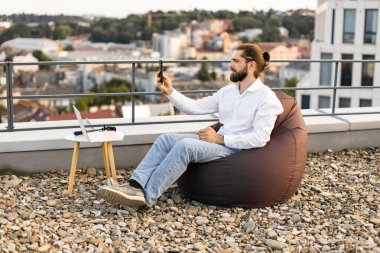 Çatıdaki adam uzaktan bilgisayar ve akıllı telefon kullanarak çalışıyor. Arka planda şehir manzarası olan günlük kıyafetler giymişti. Uzaktan çalışma, esneklik, teknoloji, açık hava ofisi, modern yaşam tarzı kavramı.