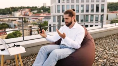 Çatıdaki adam uzaktan bilgisayar ve akıllı telefon kullanarak çalışıyor. Arka planda şehir manzarası olan günlük kıyafetler giymişti. Uzaktan çalışma, esneklik, teknoloji, açık hava ofisi, modern yaşam tarzı kavramı.