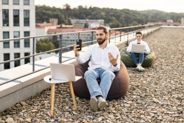 Çatıda uzaktan çalışan iş adamları, dizüstü bilgisayarlar ve akıllı telefonlar kullanıyorlar, açık ofis düzeninin tadını çıkarıyorlar. Fasulye torbaları, iletişim ve teknoloji ile şehir ortamında rahat çalışma ortamı