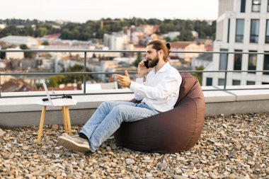 Uzaktan kumandalı fasulye torbasında çalışan bir adam küçük bir masada bilgisayarlı telefonla konuşuyor. Şehir içi rahat iş ortamı.