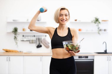 Mutfakta elinde dambıl ve bir kase sağlıklı salata tutan gülümseyen kadın. Sağlıklı yaşam tarzı, zindelik, beslenme ve sağlık kavramı.