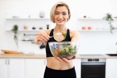 Atletizmde gülümseyen kadın modern mutfakta sağlıklı salata hazırlıyor. Sağlıklı yaşam, zindelik ve dengeli beslenmeyi teşvik eder. Cam kasede taze sebzeler..