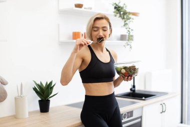 Modern mutfakta sağlıklı salatadan hoşlanan sağlıklı bir kadın atletik kıyafetler giyer. Sağlıklı yaşam tarzını, uygunluğunu ve sağlığını temsil ediyor. Parlak ve temiz ortam tazeliği ve canlılığı vurgular.