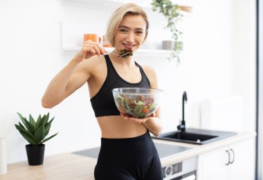 Gülümseyen kadın modern mutfakta taze salata yiyor. Sağlıklı yaşam tarzını, uygunluğunu, dengeli beslenme düzenini ve sağlığını teşvik ediyor. Canlı ve taze atmosfer sağlıklı beslenme alışkanlıkları gösteriyor..
