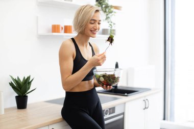 Spor giyim kuşağında gülümseyen kadın modern mutfakta taze salata yiyor. Sağlıklı beslenme, zindelik ve sağIık kavramı. Sağlıklı yaşam tarzını ve dengeli beslenmeyi seven kadınlar.