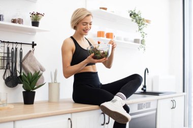 Spor giyimli formalı bir kadın tezgahta oturmuş salata yiyor. Parlak ve modern mutfak. Pozitif ve sağlıklı yaşam tarzı kavramı.
