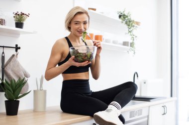 Sağlıklı salata yerken gülümseyen bir kadın. Parlak, modern mutfak. Sağlıklı yaşam tarzı, zindelik ve dengeli beslenme.