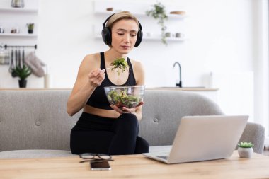 Kadın evde çalışırken taze salatanın tadını çıkarıyor. Kulaklık ve spor kıyafetlerle rahat koltukta dizüstü bilgisayarla oturmak. Sağlıklı yaşam tarzı ve uzak çalışma konsepti.