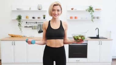Modern mutfakta elinde mavi dambıl ve bir kase salata tutan spor kıyafetli genç bir kadın. Sağlıklı yaşam tarzı, diyet ve egzersizi olumlu, neşeli bir ifadeyle destekliyor..
