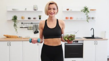 Spor kıyafetli, elinde salata kasesi ve halterle gülümseyen kadın modern mutfakta sağlıklı yaşam tarzını, diyetini ve fitnesini temsil ediyor..