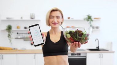 Spor giyim kuşağında elinde fitness takip uygulaması olan akıllı telefonuyla gülümseyen kadın modern mutfakta ilerleme ve bir kase salata sergiliyor. Sağlıklı yaşam tarzı, beslenme, fiziksel aktivite..