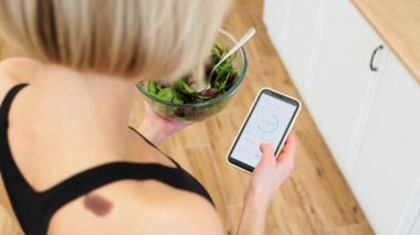 Spor elbiseli bir kadın elinde taze salata kasesiyle akıllı telefondan günlük kalori alımını takip ediyor. Sağlıklı beslenme, zindelik, beslenme ve teknoloji kavramı.