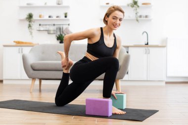 Kadın evde yoga yapıyor, bloklar kullanıyor. Parlak oturma odasında paspasın üzerinde esneme egzersizi yapıyor. Fitness, farkındalık, esneklik, rahatlama üzerine odaklanın