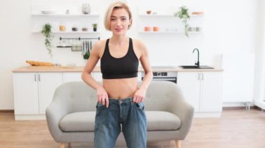 Kilo kaybettiğini gösteren bol kotları gösterirken gülümseyen bir kadın. Parlak iç mekandaki fitness başarısını kutluyoruz. Spor sutyen giyen sağlıklı bir kadın oturma odasında bitki ve raflarla dikiliyor..