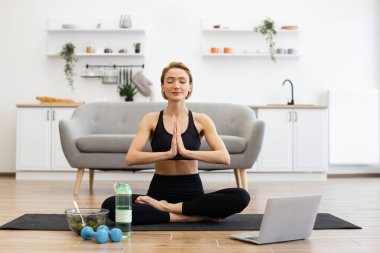 Siyah spor kıyafetli bir kadın parlak oturma odasında yoga yapıyor. Laptop, su şişesi, dambıl ve yerde salata kasesi var. Sağlık, sağlık ve farkındalığı vurgular.