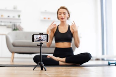 Yoga eğitmeni, akıllı telefon kullanarak paspas kaydedilen sanal fitness sınıfında bacak bacak üstüne atmış oturuyor. Eğitmen modern oturma odasında poz veriyor. Çevrimiçi fitness ve dijital sağlık kavramı.