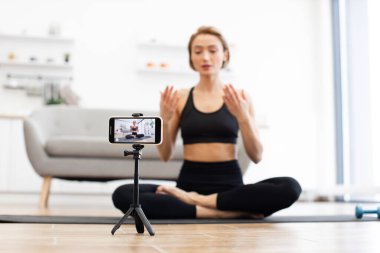Siyah spor giyim kuşağındaki kadın evden gelen tripodla yoga sınıfını kaydediyor. İç mekan fitness eğitimi, farkındalık ve modern oturma odası düzeninde tasvir edilen sağlıklı yaşam tarzı kavramları..