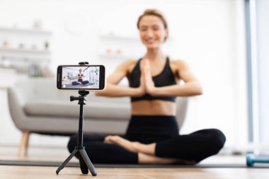 Evde yoga yapan bir kadın tripodlu akıllı telefonuyla çevrimiçi video dersi alıyor. Akıllı telefon yakalama pozisyonuna odaklan. Spor anlayışı, sağlık, ev egzersizi, sağlıklı yaşam tarzı..