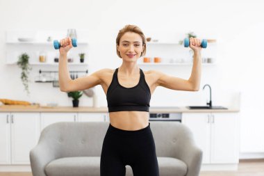 Gülümseyen kadın ev içinde egzersiz yaparken dambılları kaldırıyor. Spor giyim sektöründe bir kadın oturma odasında spor yapıyor. Sağlıklı yaşam tarzı, ev egzersizi..