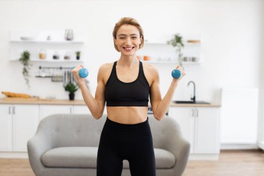 Gülümseyen kadın modern mutfakta mavi halterlerle spor yapıyor. Ev çalışması, sağlıklı yaşam tarzı ve dayanıklılık eğitimi konsepti..