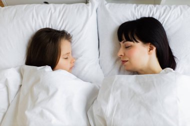 Anne ve kızı beyaz yatakta birlikte uyuyorlar. Aile üyeleri arasındaki bağı, sevgiyi ve rahatlığı yakalamak için huzurlu bir an. Huzur, masumiyet ve sıcaklığı gösterir..