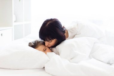 Anne, rahat beyaz yatakta uyuyan çocuğa sevgi dolu bir öpücük veriyor. Ebeveyn ve çocuk arasındaki sıcak ve sevgi dolu bir an. Şefkat ve rahatlık sahnesi