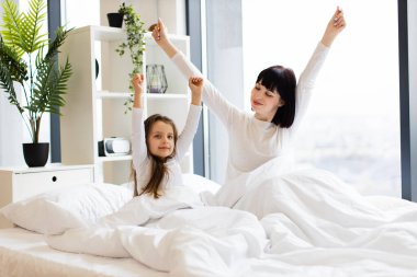 Anne ve kızı aydınlık yatak odasında sabah esnemesinin tadını çıkarıyorlar. Mutlu aile zamanları ve güne olumlu bir başlangıç. Beyaz yatak ve doğal ışık ile sıcak ev atmosferi.