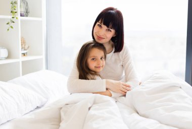 Anne ve kızı yatakta sarılıyor. Anne ve çocuk sıcak bir sabah geçiriyorlar, sevgiyi ve birlikteliği paylaşıyorlar. Parlak ve huzurlu yatak odası..