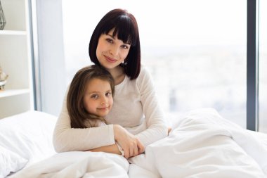 Anne ve kızı yatakta sarılıyor. Annesinin çocuğunu kucakladığı sevgi dolu bir aile anı. Ebeveyn ve çocuk arasındaki sıcak bağ ile sıcak ev atmosferi.