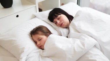 Anne ve çocuğun beyaz çarşaflarda birlikte uyuduğu huzurlu bir sahne. Sıcaklık, rahatlık ve sevgi taşıyan rahat yüzlere yakın çekim. Sıcak ve samimi aile anı.