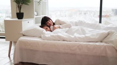 Anne ve çocuk yumuşak yastıklarla beyaz yatakta sarılıyorlar. Parlak sabah ışığı dolum odası, sıcak, rahat bir atmosfer yaratıyor. Aile bağları, sevgi, ilgi ve rahatlık taşınır.