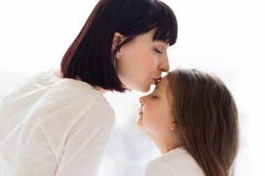 Arkadan bak. Sevgili anne kızını alnından öpüyor, şefkat, ilgi ve şefkat gösteriyor. Aile bağına, sıcaklığa, bağlantıya yakın çekim. Ebeveyn ve çocuk arasında nazik bir an. Yumuşak ışık arkaplanı.