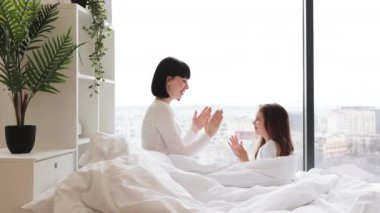 Anne ve kızı yatakta oynuyorlar. Parlak, rahat yatak odasında el çırpıyorlar. Sıcak aile anı, neşe ve bağ. Kapalı mekan sahnesi.