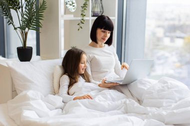 Aile bağları, teknoloji kullanımı ve rahatlama kavramı. Anne ve kızı parlak odada dizüstü bilgisayar kullanarak rahat bir yatakta uzanıyorlar. Aile bağları, teknoloji kullanımı ve rahatlama kavramı.