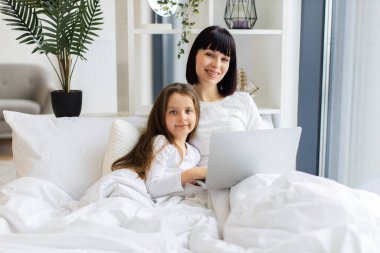 Parlak, rahat yatak odası sıcak bir aile ortamı yaratıyor. Anne ve kızı dizüstü bilgisayar kullanarak yatakta yatıyor. Zamana bağlanmak, gülümsemek ve teknolojinin tadını çıkarmak..