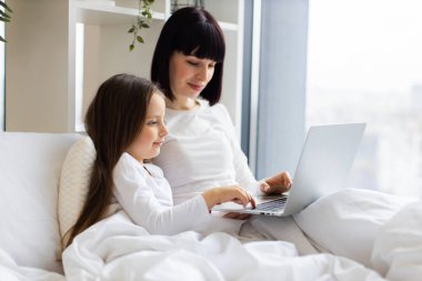 Parlak ve rahat yatak odası rahat bir atmosfer yaratıyor. Anne ve kızı dizüstü bilgisayar kullanarak yatakta yatıyor. Sıcak aile zamanı, birliktelik, teknoloji etkileşimi ve çevrimiçi aktiviteler..