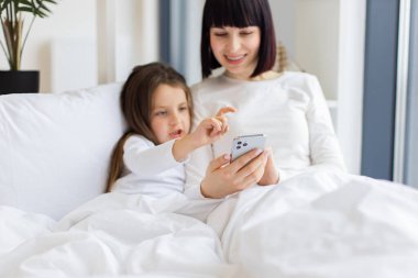 Sıcak yatak odası ortamında çekilen mutlu bir bağ anı. Anne ve kızı yatakta uzanıp akıllı telefon kullanarak eğleniyorlar. Anne ve çocuk birlikte iyi vakit geçiriyorlar..