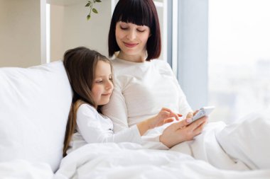 Mutlu aile bağları, anıları paylaşmak ve sıcak bir yuva ortamında birlikte olmanın tadını çıkarmak. Anne ve kızı yatakta uzanıp akıllı telefon kullanarak eğleniyorlar..