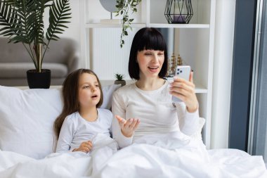 Parlak, rahat yatak odası. Anne ve kızı yatakta akıllı telefondan görüntülü aramanın keyfini çıkarıyorlar. Teknoloji ve iletişim ile aile bağı kurma anı. Mutlu ifadeler, rahat atmosfer..