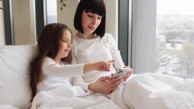 Pencereden gelen parlak doğal ışık rahat bir atmosfer sağlıyor. Anne ve kızı yatakta uzanmış telefonda gülümserken görüntülü konuşma yapıyorlar. Şehir manzarası pencerenin dışında görünür.