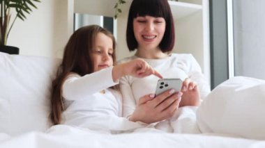 Sıcak yatak odası ortamında çekilen mutlu bir bağ anı. Anne ve kızı yatakta uzanıp akıllı telefon kullanarak eğleniyorlar. Anne ve çocuk birlikte iyi vakit geçiriyorlar..