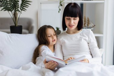 Anne yatak odasında kızıyla kitap okuyor. Aile bağı kurma, birlikte güzel vakit geçirme. Aynı beyaz kıyafetleri giyen kadın ve kız, rahat bir şekilde yatakta oturuyorlar..