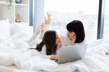 Anne ve kızı yatakta birlikte güzel vakit geçiriyorlar. İkisi de rahat kıyafetler giyiyor, dizüstü bilgisayar kullanıyor, sohbet ediyorlar. Pencereden gelen parlak doğal ışık, rahat ev ortamı.