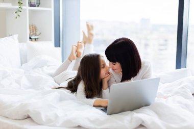 Anne ve kızı dizüstü bilgisayarla yatakta güzel vakit geçiriyorlar. Aile bağları, sevgi ve bağlar pencereden gelen parlak doğal ışıkla vurgulanır..
