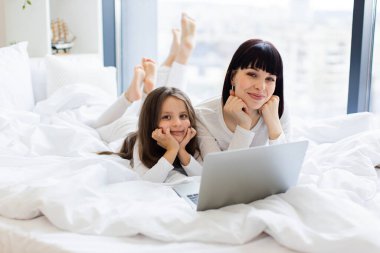 Anne ve kızı birlikte dizüstü bilgisayar kullanarak yatakta yatıyorlar. Konforlu ev ortamında mutlu aile bağları. Gülen yüzler sevgiyi, birlikteliği ve rahatlığı gösterir..
