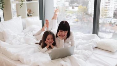 Anne ve kızı yatakta birlikte güzel vakit geçiriyorlar. İkisi de rahat kıyafetler giyiyor, dizüstü bilgisayar kullanıyor, sohbet ediyorlar. Pencereden gelen parlak doğal ışık, rahat ev ortamı.